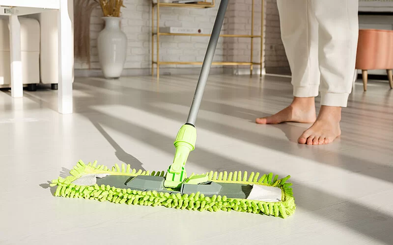 Eine Frau reinigt mit einem grünen Wischmopp einen Fußboden mit hellem Kunststoffbelag.