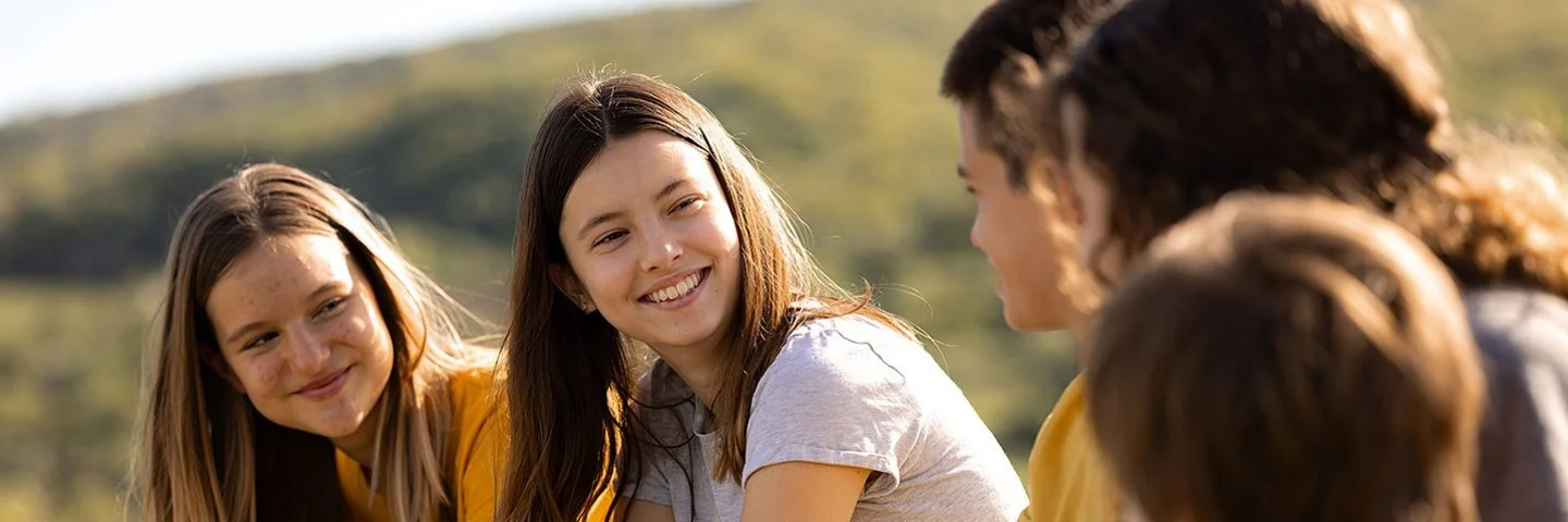 An einem sonnigen Tag sitzen fünf Jugendliche, drei Mädchen und zwei Jungen, auf einer Wiese. Die beiden Mädchen links schauen lächelnd zu den drei Kindern rechts hinüber.
