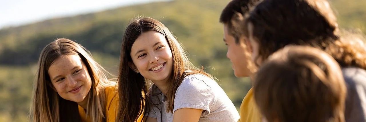 An einem sonnigen Tag sitzen fünf Jugendliche, drei Mädchen und zwei Jungen, auf einer Wiese. Die beiden Mädchen links schauen lächelnd zu den drei Kindern rechts hinüber.