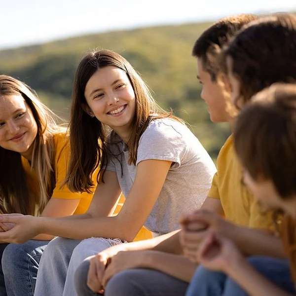 An einem sonnigen Tag sitzen fünf Jugendliche, drei Mädchen und zwei Jungen, auf einer Wiese. Die beiden Mädchen links schauen lächelnd zu den drei Kindern rechts hinüber.