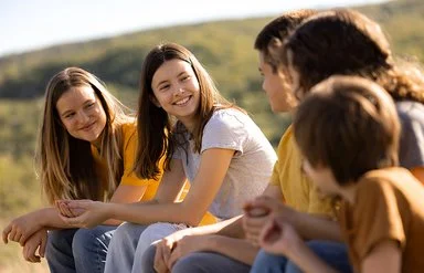 An einem sonnigen Tag sitzen fünf Jugendliche, drei Mädchen und zwei Jungen, auf einer Wiese. Die beiden Mädchen links schauen lächelnd zu den drei Kindern rechts hinüber.