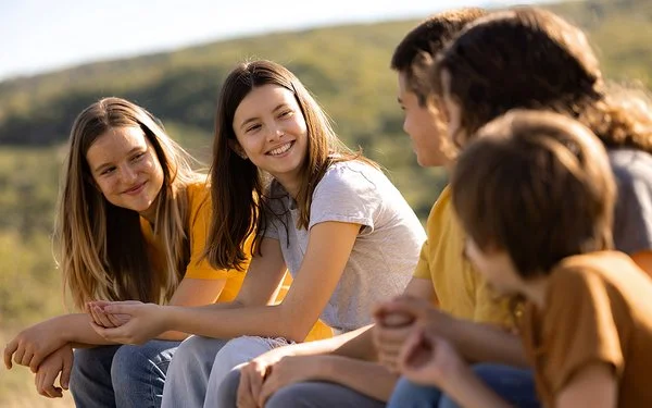 An einem sonnigen Tag sitzen fünf Jugendliche, drei Mädchen und zwei Jungen, auf einer Wiese. Die beiden Mädchen links schauen lächelnd zu den drei Kindern rechts hinüber.