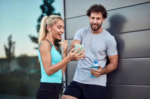 Ein Mann und eine Frau in Sportbekleidung stehen vor einer Wand und schauen auf ein Smartphone.