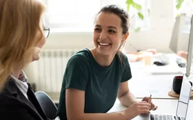 Eine jnuge Frau sitzt an einem Tisch vor einem Laptop und lächelt eine andere, rechts neben ihr sitzende Frau an.