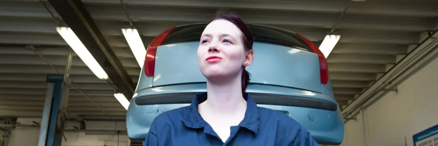 Eine junge Person in blauer Arbeitskleidung mit orangefarbenem Handschuh steht vor einem aufgebockten Auto in einer Werkstatt.