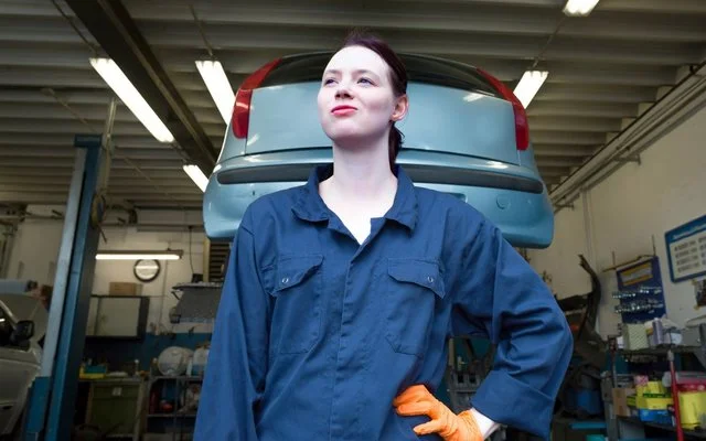 Eine junge Person in blauer Arbeitskleidung mit orangefarbenem Handschuh steht vor einem aufgebockten Auto in einer Werkstatt.