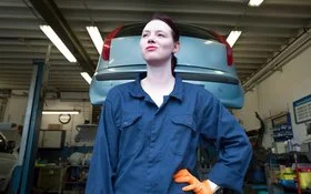 Eine junge Person in blauer Arbeitskleidung mit orangefarbenem Handschuh steht vor einem aufgebockten Auto in einer Werkstatt.