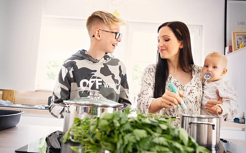 Eine Frau und ein Junge im Teenageralter in der Küche. Im Vordergrund sind frische Kräuter und Salat zu sehen. Die Frau hält einen Säugling im Arm. Sie und der Junge bereiten ein Essen zu.