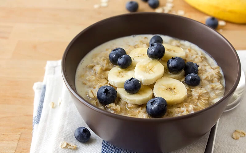 Eine kleine braune Keramikschale, appetitlich gefüllt mit Haferbrei, in Scheiben geschnittenen Bananen und ein paar Blaubeeren steht auf einem gefalteten weiß-blauen Küchenhandtuch, das auf einem hellen Holztisch liegt. Ein paar Blaubeeren und Haferflocken liegen verstreut auf dem Tisch, im Hintergrund ist eine Banane zu sehen.