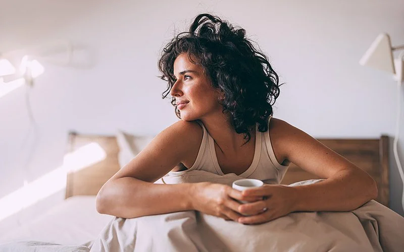 In einem hellen Schlafzimmer sitzt eine Frau auf einem Bett mit der Bettdecke über den Beinen. Sie hält einen Kaffeebecher und blickt lächelnd in die Richtung, aus der Licht ins Zimmer fällt.