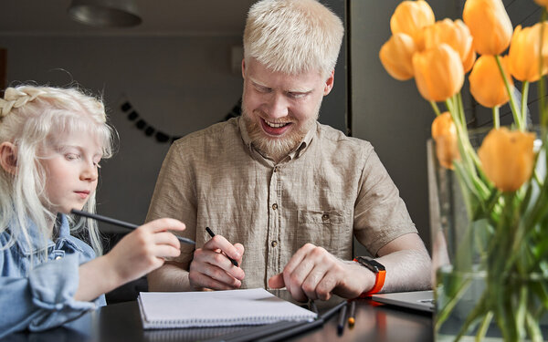 Vater mit Albinismus sitzt mit seinem Kind, welches ebenfalls Albinismus hat, am Tisch.