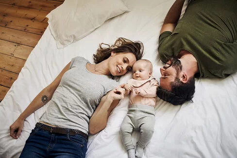 Hochwinkelaufnahme eines liebevollen jungen Paares, das mit seinem Baby auf dem Bett liegt, lächelt und spielt.