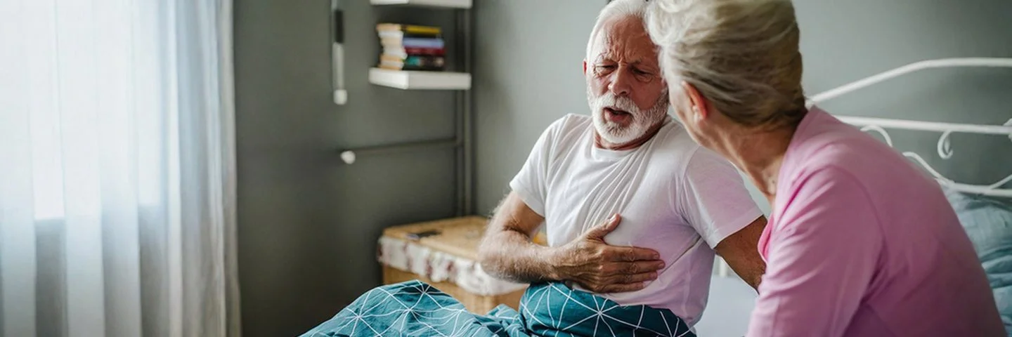 Ein senior sitzt in seinem Bett und fasst sich an die Brust. Eine ältere Frau wendet sich ihm zu.