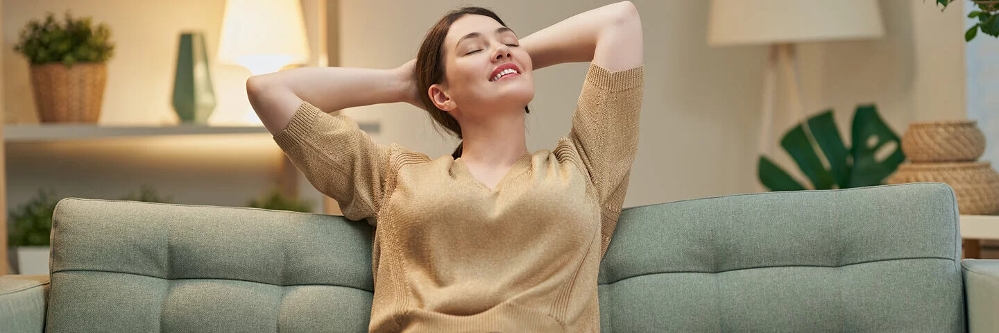 Eine Frau sitzt auf der Couch und fühlt sich glücklich und gesund in ihrem Körper.
