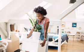 Eine fröhliche junge Frau, die gerade in ihre erste eigene Wohnung gezogen ist, steht mit Umzugskisten im Raum.