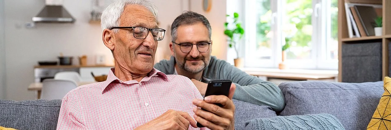 Ein alter Mann sitzt auf einem frei im Raum stehenden Sofa. Er hält ein Smartphone in der linken Hand und liest lächelnd darauf. Hinter dem Sofa kniet ein jüngerer Mann, der sich mit den Unterarmen auf der Sofalehne abstützt. Er blickt dem Älteren über die Schulter auf das Display und lächelt.