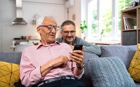 Ein alter Mann sitzt auf einem frei im Raum stehenden Sofa. Er hält ein Smartphone in der linken Hand und liest lächelnd darauf. Hinter dem Sofa kniet ein jüngerer Mann, der sich mit den Unterarmen auf der Sofalehne abstützt. Er blickt dem Älteren über die Schulter auf das Display und lächelt.