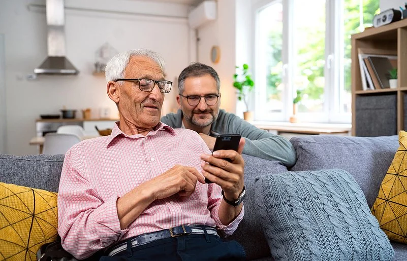 Ein alter Mann sitzt auf einem frei im Raum stehenden Sofa. Er hält ein Smartphone in der linken Hand und liest lächelnd darauf. Hinter dem Sofa kniet ein jüngerer Mann, der sich mit den Unterarmen auf der Sofalehne abstützt. Er blickt dem Älteren über die Schulter auf das Display und lächelt.