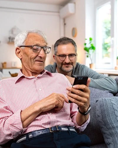 Ein alter Mann sitzt auf einem frei im Raum stehenden Sofa. Er hält ein Smartphone in der linken Hand und liest lächelnd darauf. Hinter dem Sofa kniet ein jüngerer Mann, der sich mit den Unterarmen auf der Sofalehne abstützt. Er blickt dem Älteren über die Schulter auf das Display und lächelt.