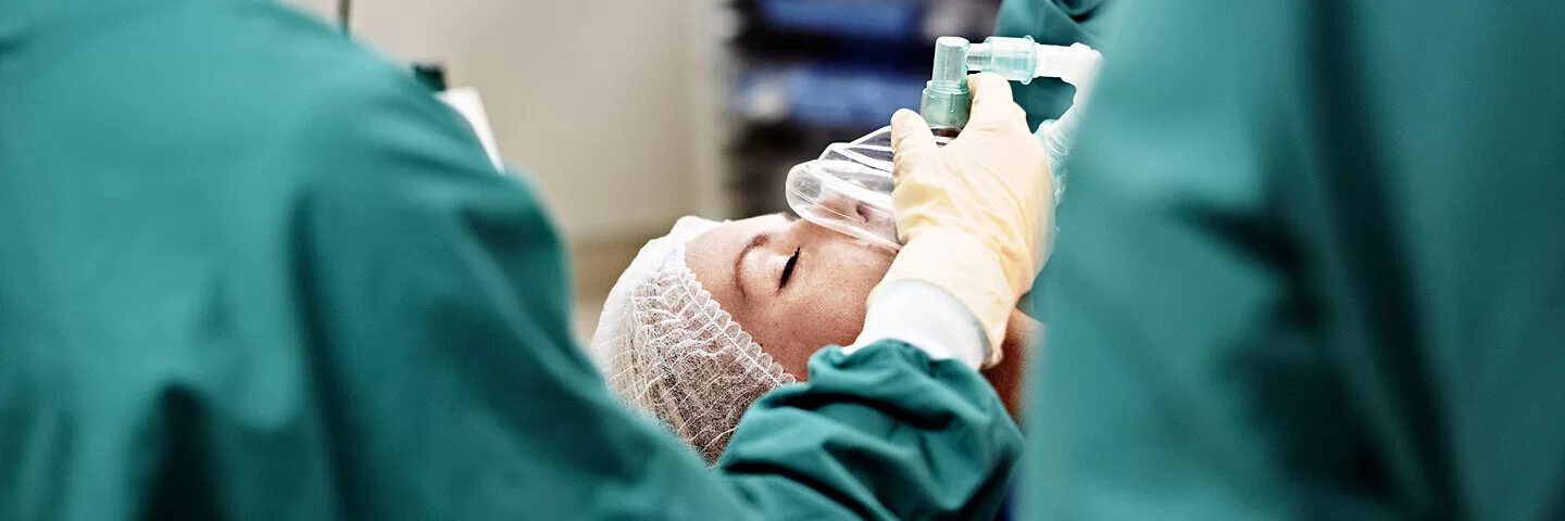 An einem OP-Tisch gibt ein Anästhesist über eine Atemmaske einer Patientin Sauerstoff.