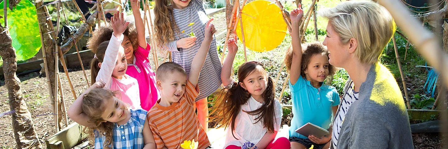 Eine Lehrerin mit rot-blonden Haaren, blauer Bluse und einem Tablet in den Händen lächelt in die Kamera. Im Hintergrund ist ein Klassenzimmer mit Kindern zu sehen, die an ihren Plätzen arbeiten.