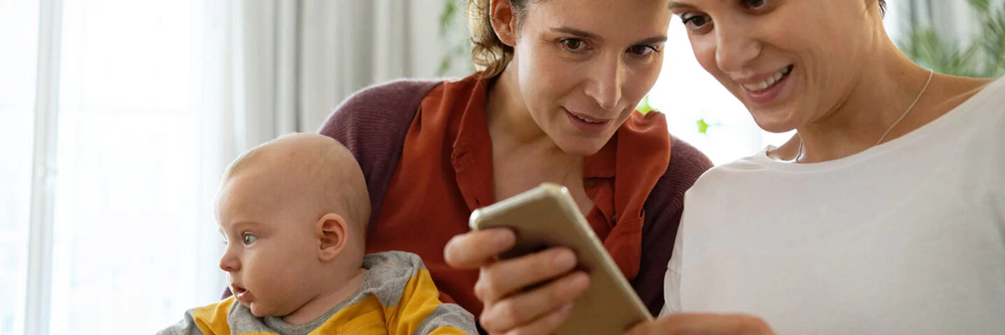Zwei Frauen sitzen in einem Zimmer und schauen auf ein Handy. Eine der beiden hält ein Baby im Arm.