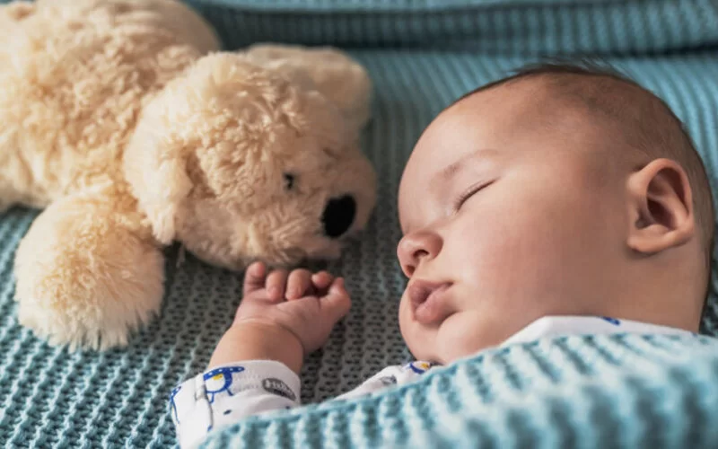 Ein Baby schläft auf einer Decke mit einem Teddybären im Hintergrund.
