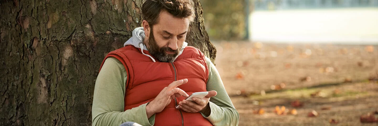 Ein Mann sitzt im Park auf der Wiese und lehnt sich an einem Baum an. In der Hand hält er ein Smartphone und gibt etwas darauf ein.