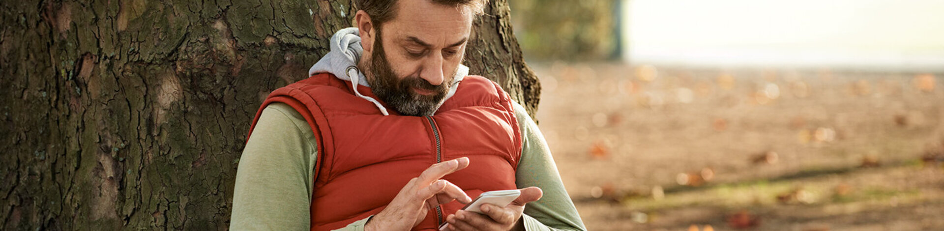 Ein Mann sitzt im Park auf der Wiese und lehnt sich an einem Baum an. In der Hand hält er ein Smartphone und gibt etwas darauf ein.