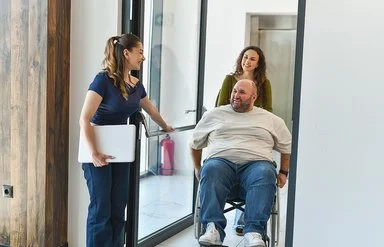 Eine junge Frau hält einem Mann, der im Rollstuhl sitzt und von einer anderen Frau geschoben wird, eine große Glastür auf.