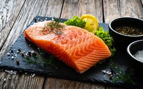 Ein Stück Lachs liegt auf einer Schieferplatte, daneben Gewürze, Salat und eine Scheibe Zitrone.