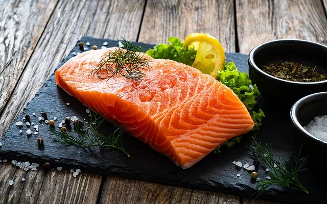 Ein Stück Lachs liegt auf einer Schieferplatte, daneben Gewürze, Salat und eine Scheibe Zitrone.