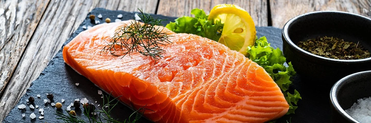 Ein Stück Lachs liegt auf einer Schieferplatte, daneben Gewürze, Salat und eine Scheibe Zitrone.