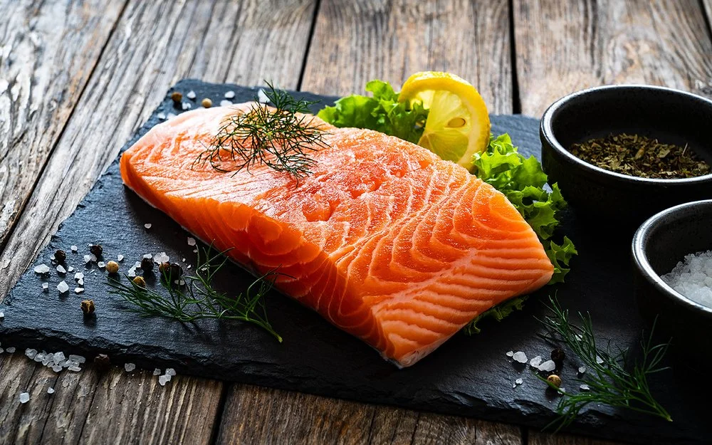 Ein Stück Lachs liegt auf einer Schieferplatte, daneben Gewürze, Salat und eine Scheibe Zitrone.