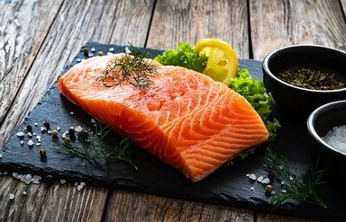 Ein Stück Lachs liegt auf einer Schieferplatte, daneben Gewürze, Salat und eine Scheibe Zitrone.