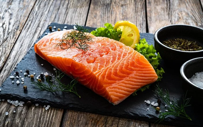 Ein Stück Lachs liegt auf einer Schieferplatte, daneben Gewürze, Salat und eine Scheibe Zitrone.