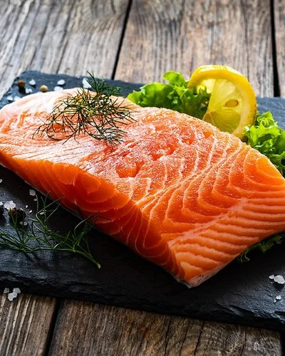 Ein Stück Lachs liegt auf einer Schieferplatte, daneben Gewürze, Salat und eine Scheibe Zitrone.