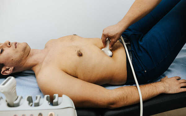 Ein junger Mann mit Leberzirrhose liegt auf einer Behandlungsliege und wird von einem Arzt oder einer Ärztin mit dem Ultraschallgerät untersucht.