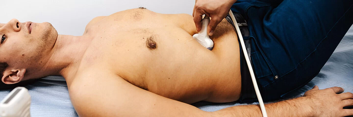 Ein junger Mann liegt auf einer Behandlungsliege und wird von einem Arzt oder einer Ärztin mit dem Ultraschallgerät untersucht.