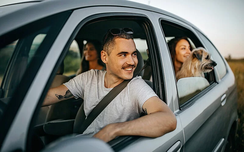 Am Steuer eines Autos sitzt ein Mann, auf dem Rücksitz zwei Frauen und ein Hund. Alle Fenster sind heruntergekurbelt.