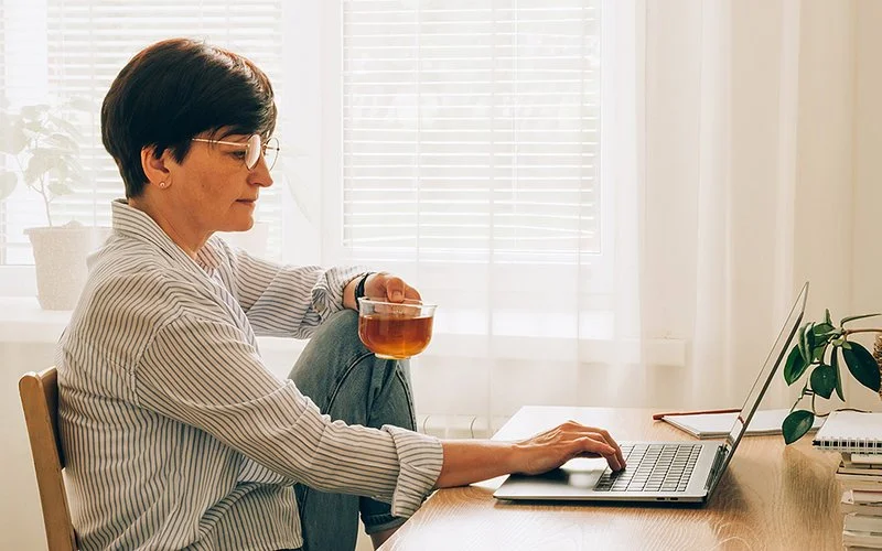 Eine Frau sitzt mit einem Teeglas in der Hand auf einem Stuhl und sieht auf einen Laptop.