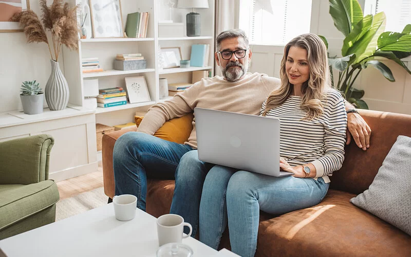 Ein Paar sitzt auf dem Sofa am Laptop und ruft den AOK Familiencoach Pflege auf.