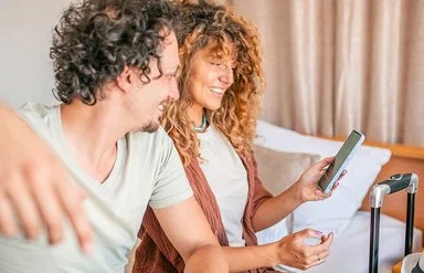 Ein junges Paar sitzt zusammen auf einem Hotelbett mit ihren Koffern und sehen auf den Bildschirm eines Handys.
