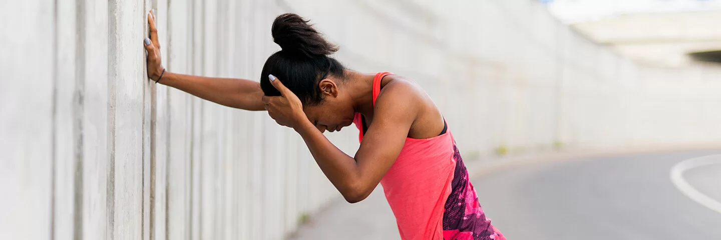 Eine Joggerin steht an einer Wand und hält sich wegen Kopfschmerzen den Kopf.