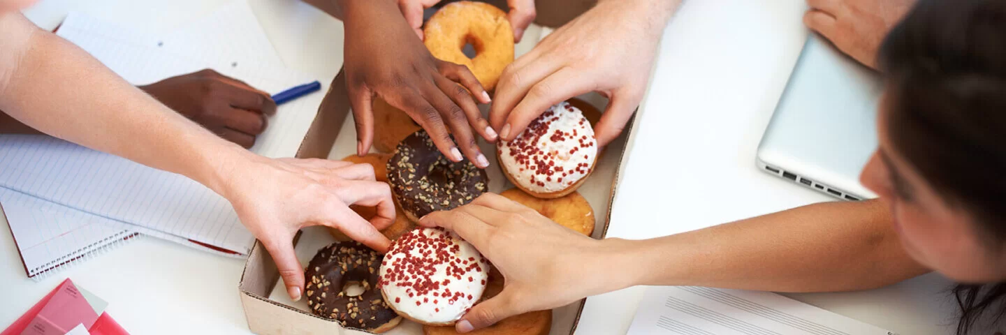 Mehrere Personen greifen nach zuckerhaltigen Donuts aus einer Schachtel bei einem Meeting.