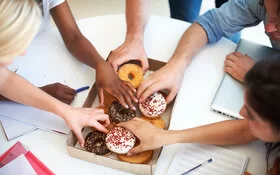 Mehrere Personen greifen nach zuckerhaltigen Donuts aus einer Schachtel bei einem Meeting.