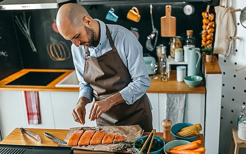 Ein Mann mit einer braunen Schürze in einer Küche würzt Lachsstücke, die auf einem Holzbrett liegen.