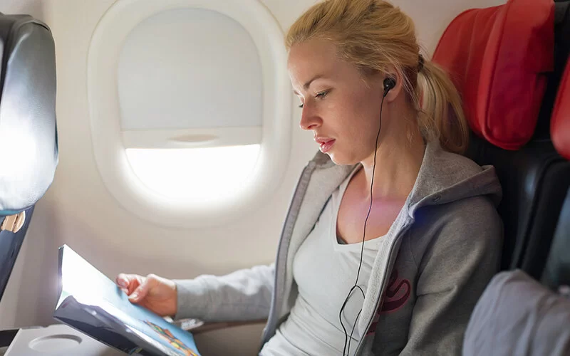 Junge Frau sitzt im Flugzeug. Sie liest eine Zeitschrift und hört dabei Musik mit Kopfhörern.