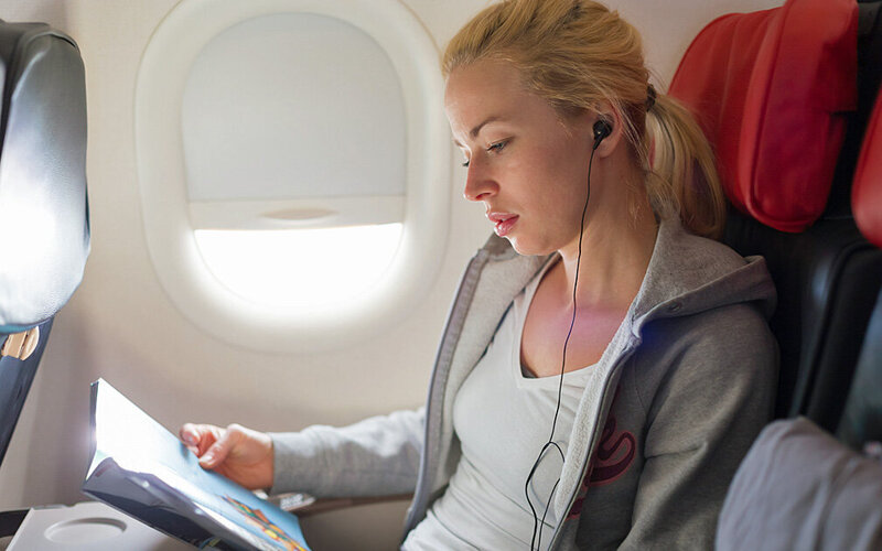 Junge Frau sitzt im Flugzeug. Sie liest eine Zeitschrift und hört dabei Musik mit Kopfhörern.