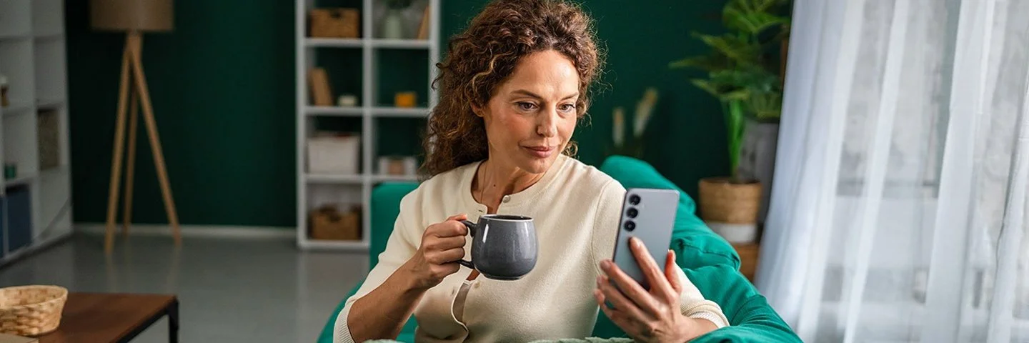 Eine Frau sitzt in einem Wohnzimmer auf einer grünen Couch. In der rechten Hand hält sie eine Tasse, in der linken Hand ihr Smartphone, auf dem sie etwas liest. Im Hintergrund sind Einrichtungsgegenstände und eine Zimmerpflanze zu sehen.